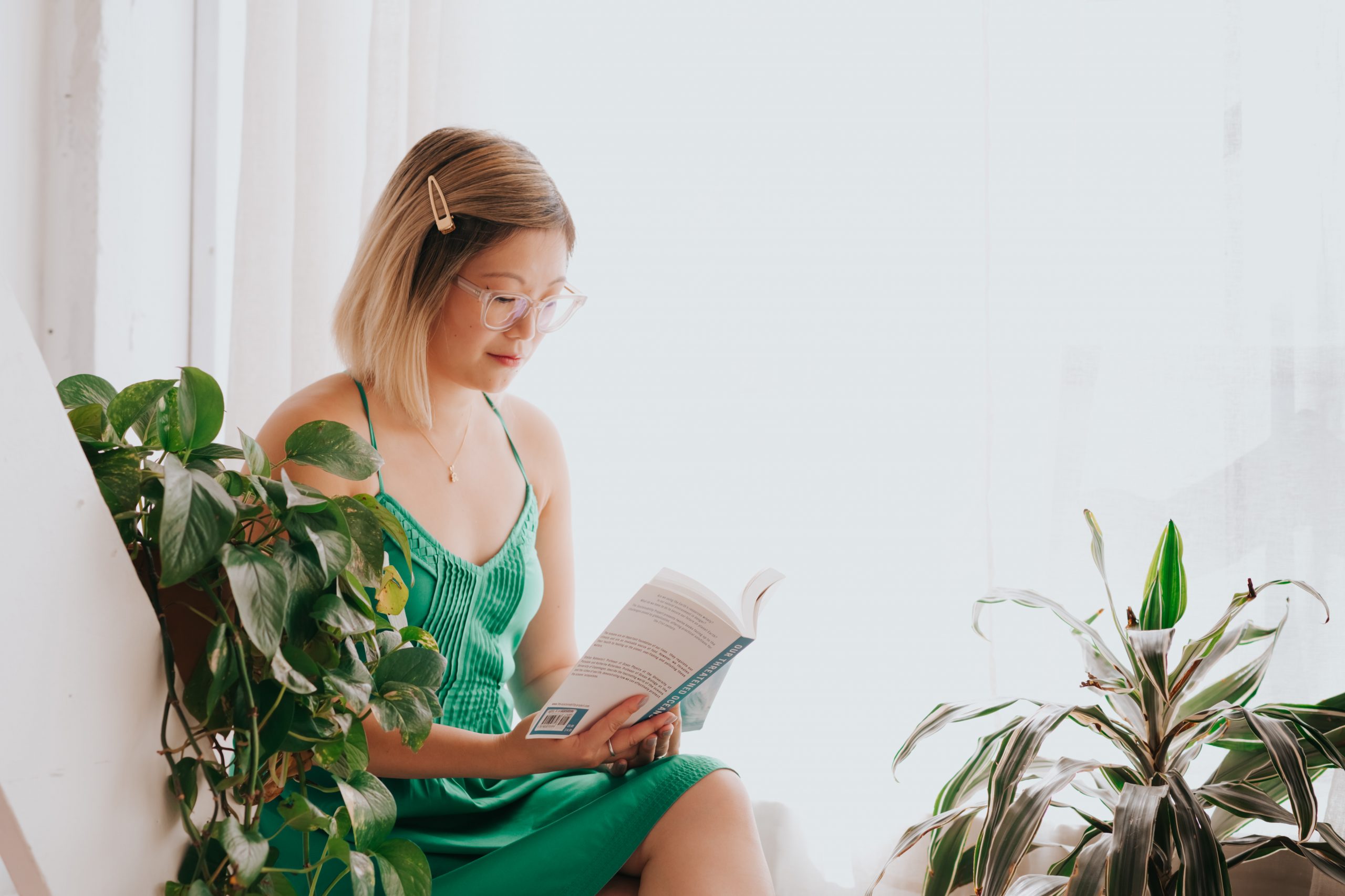 Woman in a green dress is sitting down and reading a book surrounded by plants